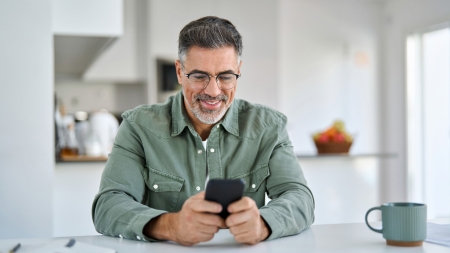 Ein Mann mit grauem Haar und Brille, der ein grünes Hemd mit Knöpfen trägt, sitzt an einem weissen Tisch in einer hellen Küche und lächelt, während er auf sein Smartphone schaut