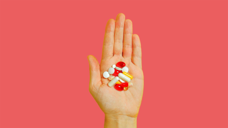 Une main ouverte tenant diverses pilules et capsules de différentes tailles et couleurs, sur un fond rose uni.