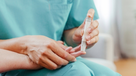 Eine medizinische Fachkraft in blaugrüner OP-Kleidung misst das Blut eines Patienten mit einem Blutzuckermessgerät. Die Hand des Patienten ruht während der Messung auf dem Schoss der Fachkraft.