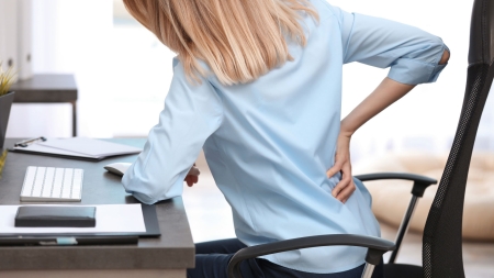 Une personne aux longs cheveux blonds, vêtue d'une chemise bleu clair, est assise à un bureau, se tourne légèrement et se tient le bas du dos d'une main, semblant être mal à l'aise. Le bureau est équipé d'un clavier, d'un bloc-notes et de papiers.