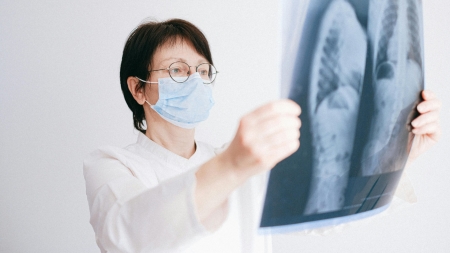 A person wearing a face mask and goggles is looking at an X-ray image in a bright room. They appear focused and attentive and are wearing a white lab coat.