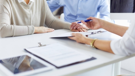 Two people are sitting at a desk, one is holding a pen above a document and they are discussing something. A tablet and a clipboard are lying on the table. The atmosphere is reminiscent of a business meeting or a consultation.