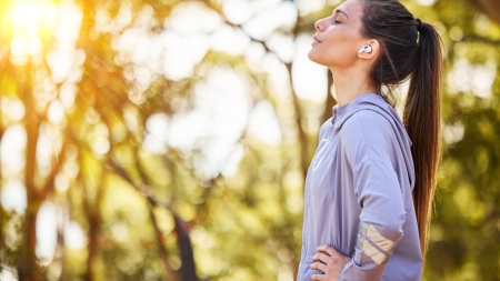 A woman with long hair tied back is standing outside. She is wearing wireless headphones and a grey hoodie. Her eyes are closed and her face is turned towards the sun. In the background, there are blurred, sunlit trees.