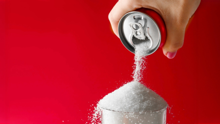 Eine Hand hält eine Getränkedose und schüttet vor einem leuchtend roten Hintergrund eine große Menge Zucker in ein Glas, um den hohen Zuckergehalt in Erfrischungsgetränken hervorzuheben.