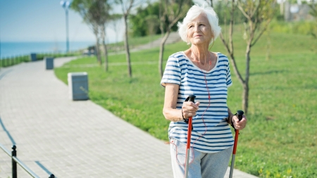 Eine ältere Frau mit weißen Haaren geht im Freien auf einem gepflasterten Weg mit Nordic-Walking-Stöcken. Sie trägt ein gestreiftes Hemd, eine graue Hose und Kopfhörer, mit grünem Gras und Bäumen im Hintergrund an einem sonnigen Tag.