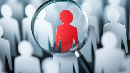 Une loupe focalise l'attention sur une silhouette humaine rouge au milieu de nombreuses silhouettes blanches découpées, symbolisant le concept de se démarquer ou d'être choisi parmi une foule.