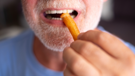 A close-up of an older man with a white beard and mustache about to eat a French fry, holding it near his open mouth.