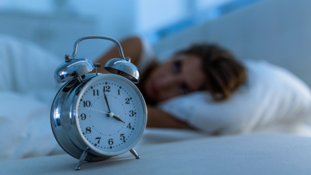 Una sveglia argentata segna le 3:00, mentre sullo sfondo una persona giace sveglia nel letto, con la testa appoggiata su un cuscino bianco, e guarda l'orologio in una stanza debolmente illuminata.