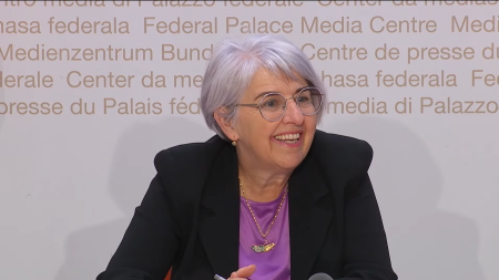 Une femme souriante aux cheveux gris courts et portant des lunettes est assise devant un fond avec un texte multilingue. Elle porte un blazer noir et une chemise violette.