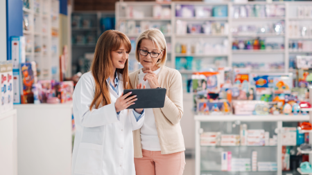 Un pharmacien en blouse blanche montre un comprimé à une femme âgée portant des lunettes dans une pharmacie bien éclairée, avec des étagères remplies de médicaments et de produits visibles en arrière-plan.