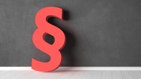 Ein großes rotes Paragraphen-Symbol (§) steht auf einem Holzboden vor einer dunkelgrauen Kreidetafelwand.