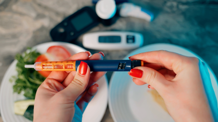 Eine Person mit rotem Nagellack hält einen Insulinstift, während auf einem Tisch im Hintergrund ein Blutzuckermessgerät, Teststreifen und Teller mit Lebensmitteln zu sehen sind.