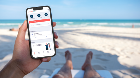 Eine Person entspannt sich auf einem Strandstuhl, ihre sandigen Füße sind zu sehen, und hält ein Smartphone in der Hand, auf dessen Bildschirm die Benutzeroberfläche einer App zu sehen ist. Das Meer und der blaue Himmel bilden den Hintergrund und schaffen eine friedliche, tropische Atmosphäre. (Symbolbild: Adobe Stock)