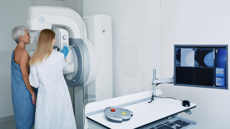 Eine medizinische Fachkraft bedient ein Mammographiegerät für eine Frau im Arztkittel und bereitet sie auf eine Brustuntersuchung vor. Ein Computermonitor in der Nähe zeigt Mammographie-Bilder an.