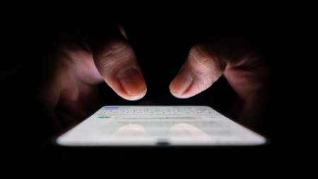 Gros plan sur des mains tapant sur un smartphone dans l'obscurité, l'écran étant allumé et éclairant les doigts. Le téléphone montre/voit une application de messagerie ou d'envoi de SMS.