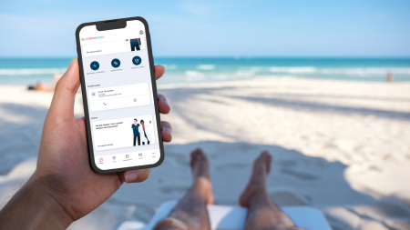Una persona si rilassa su una sedia a sdraio, con i piedi ricoperti di sabbia in primo piano, mentre tiene in mano uno smartphone sul cui schermo è visibile l'interfaccia di un'app. Il mare e il cielo azzurro fanno da sfondo, creando un'atmosfera tranquilla e tropicale.