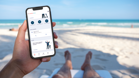 Une personne se détend sur une chaise longue, les pieds couverts de sable, un smartphone à la main dont l'écran affiche l'interface d'une application. La mer et le ciel bleu servent de toile de fond et créent une atmosphère paisible et tropicale. 