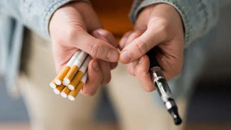 A person holds several cigarettes in one hand and a vape device in the other, suggesting a choice between traditional smoking and vaping. The image is focused on the hands and items, with a blurred background.