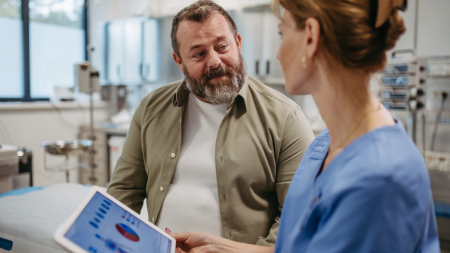 Un professionnel de la santé en blouse montre une tablette avec des graphiques à un homme en tenue décontractée lors d'une consultation dans un cabinet médical. Ils discutent, l'équipement médical étant visible à l'arrière-plan.