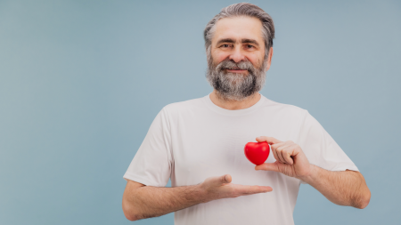 Un uomo anziano con i capelli grigi e la barba sorride mentre tiene un piccolo cuore rosso in una mano e con l'altra si avvicina ad esso.